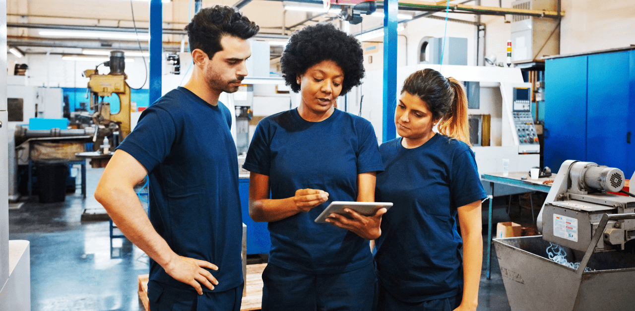 In a factory, 2 men wear blue work shirts and safety glasses as they lean on a table covered with a clipboard, cup and other items. To the right of the men, a woman wearing a grey business suit and safety glasses shows the men something on a computer. We are unable to see the computer screen. She holds protective headphones in her right hand.
