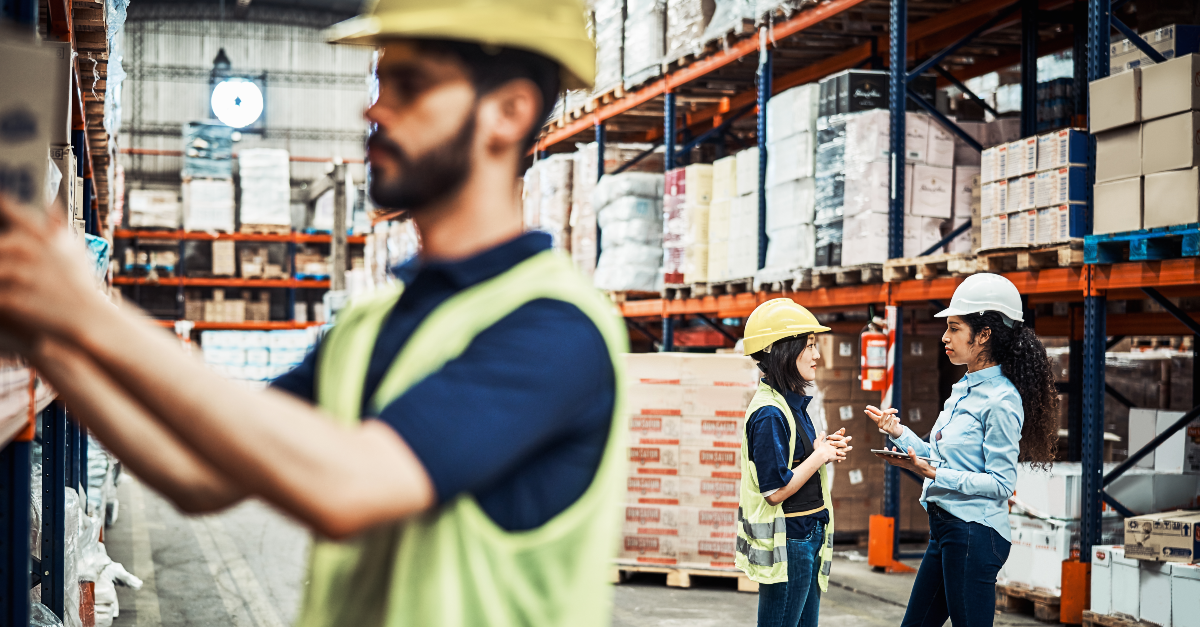 Distribution warehouse workers, a man on the left and woman on the right, move boxes onto a hand cart inside a warehouse. Both workers wear blue shirts and dark pants.