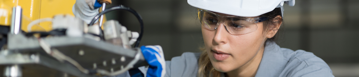 A woman engineer wearing a white hard hat and safety glasses is working on the maintenance of an autonomous robotics arm. Her correct use of PPE helps to avoid various types of OSHA violations.