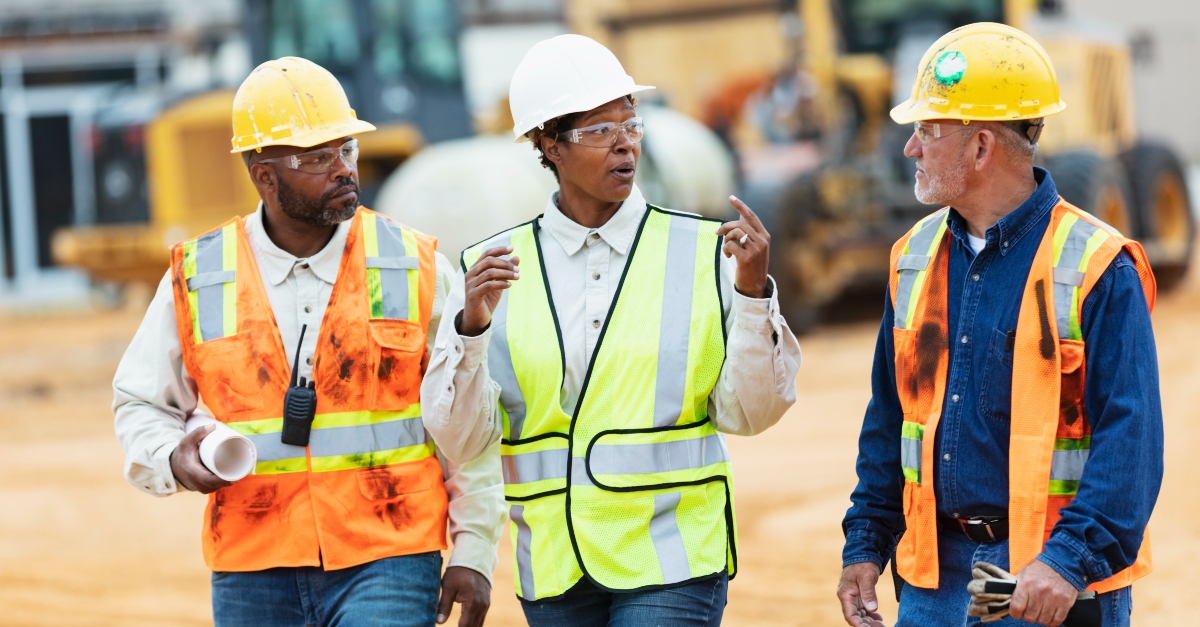 Two construction workers and a staffing agency account manager wear safety helmets and reflective vests as they tour a construction site. They are standing in front of several pieces of heavy construction equipment and vehicles. The group appear to be discussing the progress of the project. This article promotes how construction companies can benefit from the help of building and construction recruitment agencies.