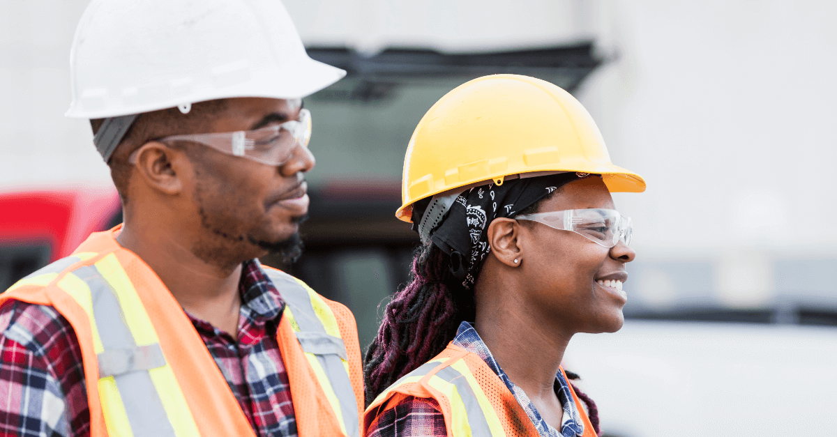 On a construction site, a woman smiles while wearing an orange safety hat, safety glasses and a high visibility vest over a flannel shirt. She is walking next to a man wearing a white hard hat, safety glasses and a high visibility vest over a flannel shirt.