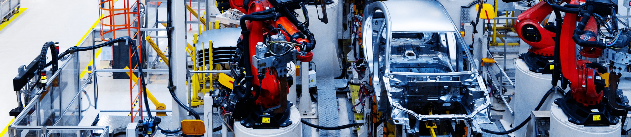 A panoramic view of a modern automotive assembly line. The scene is dominated by robotic arms in red and black, which are engaged in the meticulous assembly of a car. The car’s metallic frame shines under the facility’s bright lights, highlighting its partially assembled state and intricate internal structure. The workspace is equipped with various tools and equipment, all marked with yellow safety colors to ensure precision and efficiency. The floor is organized with white safety lines, and metal structures provide stability to the robotic arms during the assembly process