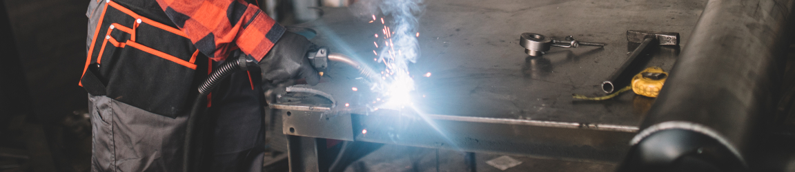 A skilled welder in safety gear is meticulously welding a metal piece on a workbench, surrounded by an array of tools and equipment indicative of a bustling workshop environment.