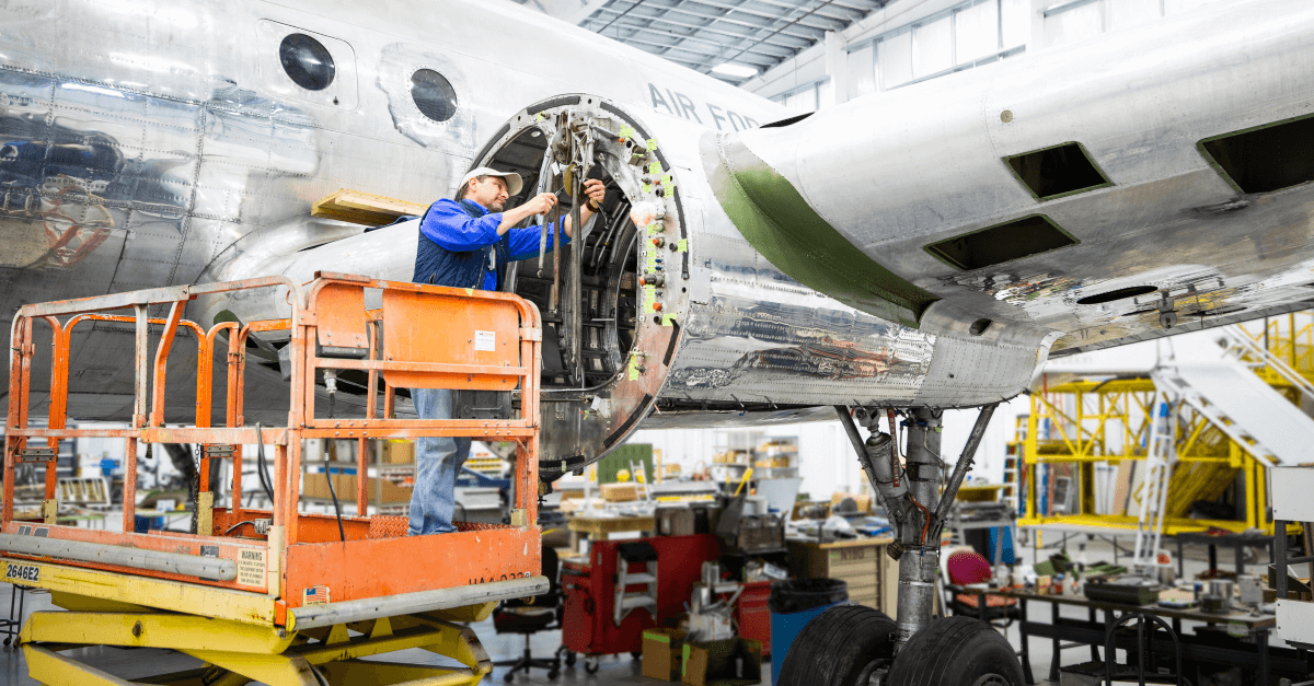 A Caucasian male aviation mechanic on a mechanical lift, works on an airplane engine in an airplane hangar. He wears a blue shirt, dark vest and white baseball cap.