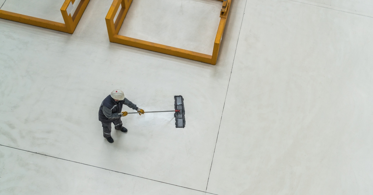 An aerial shot of a custodian cleaning the floor of a factory. The custodian is wearing a dark grey outfit and white safety hat while using a push broom.